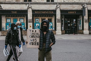 Activist of color stands outside Westminster Abbey holding a sign reading “The social contract was broken”