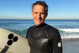 Man in a wetsuit holding a surf board in front of the ocean on a sunny day