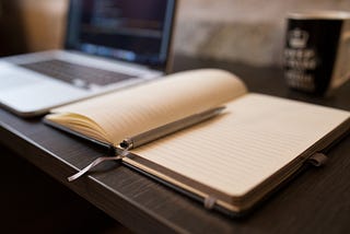 An ink pen sits atop an open journal with no words written. The journal is on a desk. A computer and mug in the background.