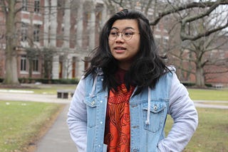 Me, a 20 year old Asian American woman with round glasses, in a sweater and a hoodie outside on my college academic quad.