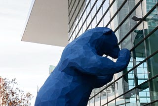 Large outside blue bear sculpture peering into the glass of the front of the Denver convention center. There are trees and building in the background.