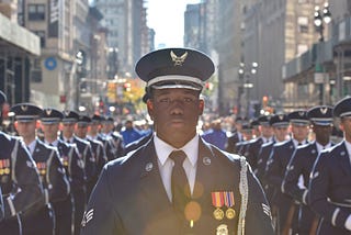 A young man with dark brown skin wears a United States Air Force uniform and stands in the streets of New York City flanked by fellow airmen for the Veterans Day Parade in 2018.