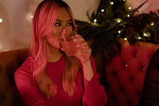 A transfemme person having a drink with christmas lights in the background.