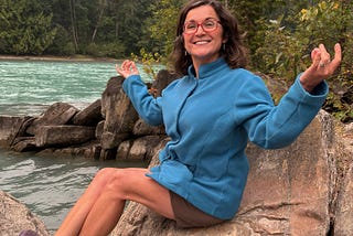 A smiling woman sitting on a boulder near a river with her arms out in a meditative position.