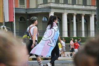 Two people walking along street, one with transgender flag being worn as a cape.