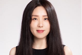 A professional headshot of a woman with long, dark hair, wearing a black sleeveless top. She is smiling slightly and looking at the camera against a plain, light-grey background.