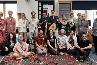 A photo of 18 people, some standing, some sitting, smiling at the camera. Some are holding instruments including lute, sitar, cello and recorder