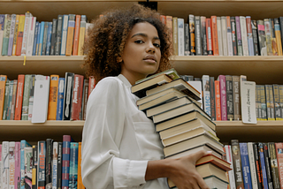 A woman standing in front of a bookshelf carrying a stack of books.