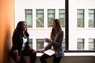 Two young women sitting in an office by the window talking and smiling