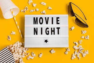 White clipboard with the words MOVIE NIGHT surrounded by popcorn, sunglasses and a drink cup. All on a bright yellow background