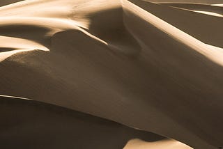 Photo by @finding_dan on Unsplash of a man walking in a desert surrounded by dunes with high contrast of light from the setting sun. A pair of footprints follow behind him.