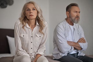 A divided mature couple sit on either side of bed with disappointed looks.