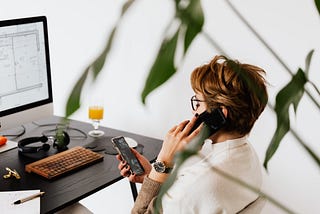 A woman sits in front of a computer. She is on the phone while looking at another one in her hand.