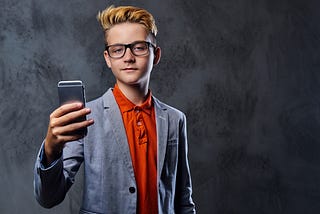 a smug-looking blond boy with glasses wearing a blazer holds up his cell phone while looking in to the camera