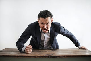 White man with angry facial expression, pounding on a wooden desk with his fist.