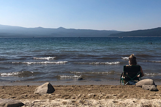 Girl sitting in a low profile camp chair along the shoreline of a lake with mountains in the distance
