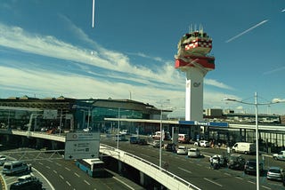 Airport seen from a hallway leading to it. The sky is clear apart from a couple of clouds and you can see the entrance to one of the terminals.