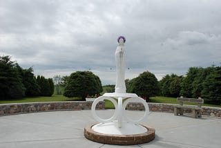 Our Lady of the Highway shrine in Vegreville. A slim white statue of a woman with trees in the background.