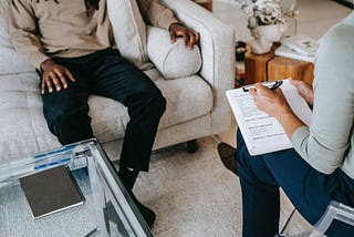 Two people, photographed from the chest down, sitting across from each other; one, facing the camera is settled into a couch, the other is sitting in a chair with a mental health exercise or assessment in their lap, taking notes.