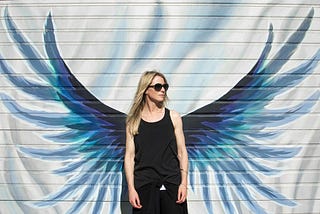 An androgynous person with long, toned arms, long, blonde hair, and dark glasses, wearing a black vest and trousers, and dark glasses, standing in front of a mural of angel wings.