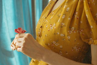 A cropped image of a woman wearing a mustard-colored, flowered dress holding a small red flower in her hands in front of her chest.