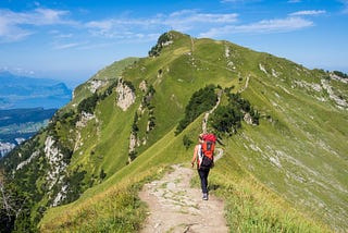 A woman with a red backpack going up a sinuous dirt mountain path, with more people higher above. The sides of the mountain are covered in grass and the clear blue sky is in the background.