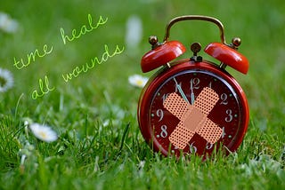 Image of an old-fashioned red alarm clock on the grass with two plasters stuck to it in a cross on the front, and to the left of it are the words: time heals all wounds.