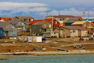 Houses sit along a shoreline. A blue sky with white clouds float overhead.