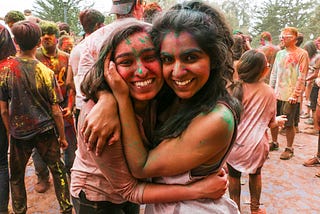 Two UC Berkeley students covered in colorful powder embrace and smile during the 2023 Holi Festival on campus.