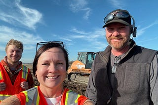 three people stand in a field with construction equipment in the background