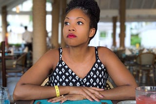 Black woman sitting at an empty dining space staring thoughtfully into the distance.