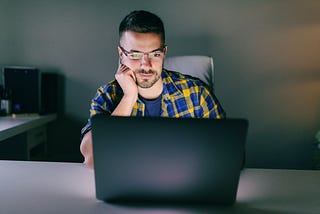 A man with glasses sitting in front of a laptop and looking concerned.