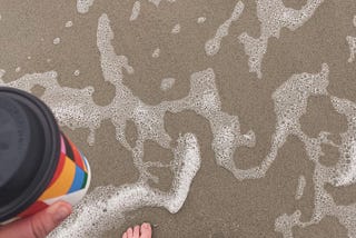 The sand and seafoam with a foot and hand holding a coffee cup.