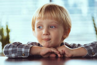 ChatGPT said: A young boy with light blond hair rests his chin on his folded hands at a table. He’s wearing a plaid shirt and looking slightly to the side with a thoughtful, almost mischievous expression. Soft daylight filters through a window behind him, giving the background a gentle blur.