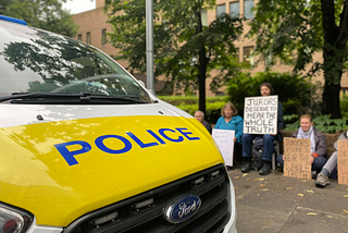 Police van sits outside crown court, people hold placards in background saying ‘jurors deserve to hear the whole truth’