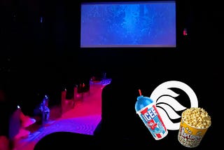 A dark theater with pink and purple flourescent floor lights. A dark blue forest scene on the screen. NYU Local logo sits with an Icee and popcorn.