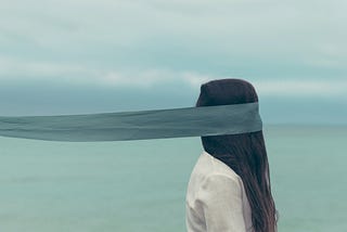 A woman standing by the ocean, a veil in front of her eyes