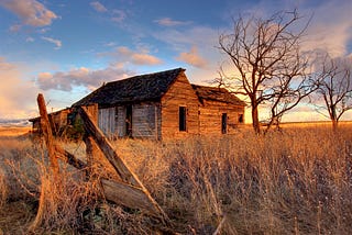 What looks like an old, rundown homestead house with aging wood siding and missing windows, in a field of grass next to a bare tree and against a partly cloudy but sunlit sky.
