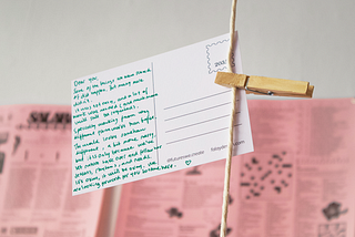 A photograph of the back of a handwritten postcard “from the future”. The postcard is attached to a string hanging from the ceiling, connected by a wooden peg.
