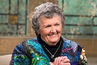 Prolific author and Benedictine Sister Joan Chittister, smiling and seated with hands clasped, before some stained glass windows.