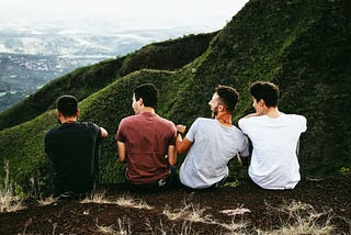 Good looking white men with brown hair sitting in a beautiful landscape of with their backs to us facing a grassy mountain having a wonderful, funny moment together, probably talking about women’s body parts.