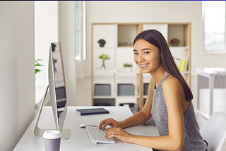 Young woman sitting at her desk, smiling at the camera, on her computer