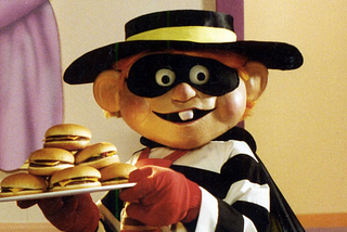 the Hamburglar is a costumed mascot of McDonald’s, wearing a hat, face mask, cape, striped top, and red gloves