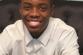 Isaiah smiling while sitting on a dark brown couch with a white short sleeved collared shirt.