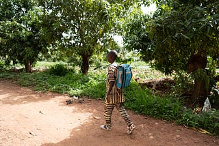 A young learner carrying a backpack walks on an unpaved road