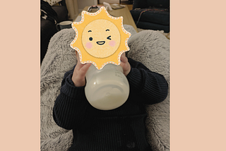 a baby drinks breast milk from a bottle. her face is obscured by a sun cartoon