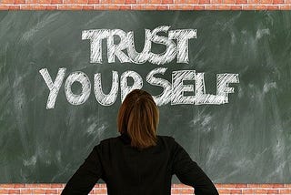 Blackboard with phrase, “Trust Yourself” written and person standing in front of it reading the text