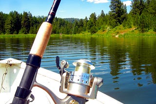 A photograph of a the handle of a fishing pole with a reel in the forground with the bow of the boat just behind in a lake looking at the shorline in the background on a sunny day with blue sky.