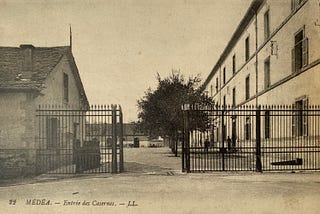 Black and white photograph of the military barracks in Medea, a three-story white building with an iron gate and a guard house.