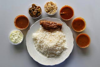 A plate with steamed rice and cooked fish placed in the center. Around the plate, seven bowls of various items like yogurt, stir-fried veggies, and curries are placed.